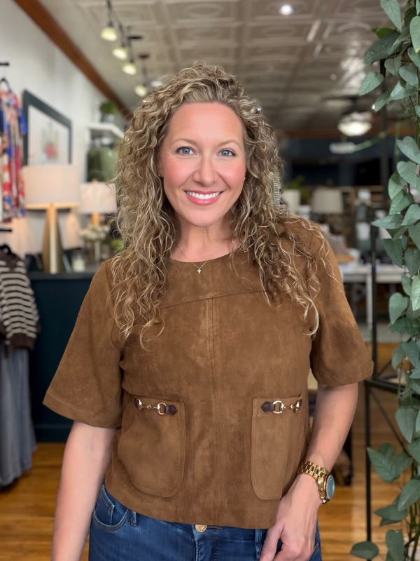 A smiling woman with curly blonde hair wearing a brown short-sleeve suede-like top with front pockets and metal accents, standing in a retail store interior with plants and clothing visible in the background.