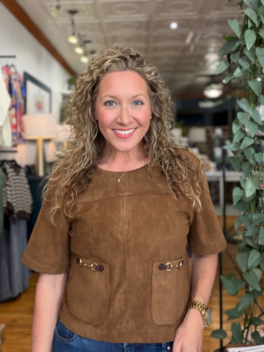 A smiling woman with curly blonde hair wearing a brown suede short-sleeve top with decorative metal accents on the front pockets, standing inside a retail store.