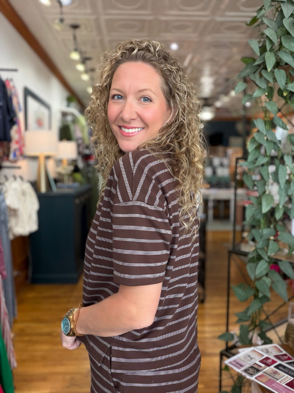 A woman with curly blonde hair wearing a brown and gray striped short-sleeve top, smiling and looking back inside a boutique store.