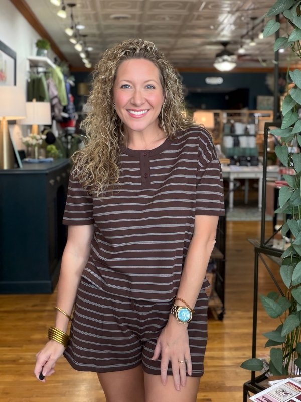 A smiling woman with curly blonde hair stands in a boutique wearing a short-sleeve brown and white striped romper. She is wearing gold bracelets and a watch, and the background shows a cozy, wood-floored shop interior with plants and clothing racks.