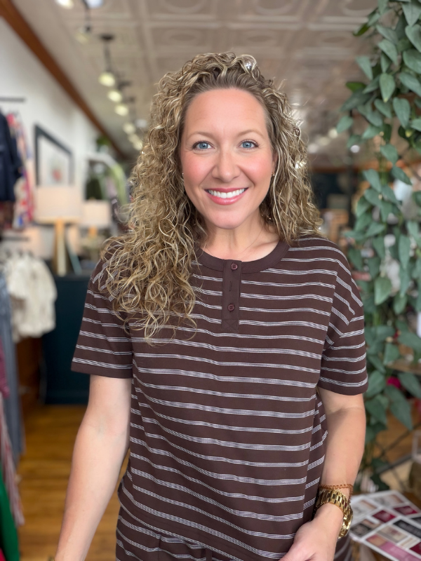 A smiling woman with curly blonde hair wearing a brown and gray horizontally striped short-sleeve henley shirt, standing inside a retail store.