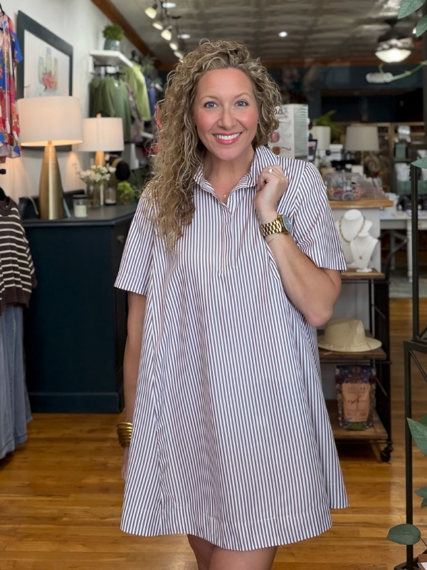 A smiling woman with curly blonde hair stands in a boutique wearing a short-sleeve button-up shirt dress with thin vertical navy and white stripes.
