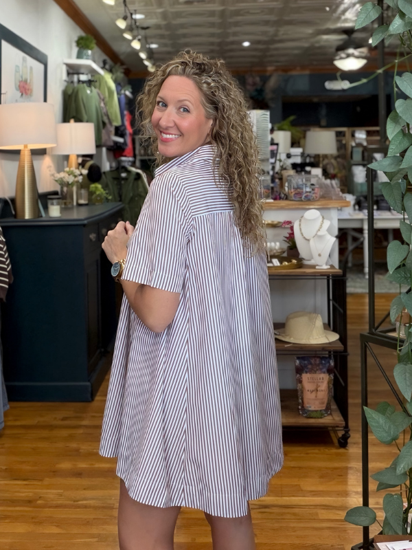 A woman with curly hair wearing a short-sleeve button-up shirt dress with thin vertical blue and white stripes, standing in a retail store.