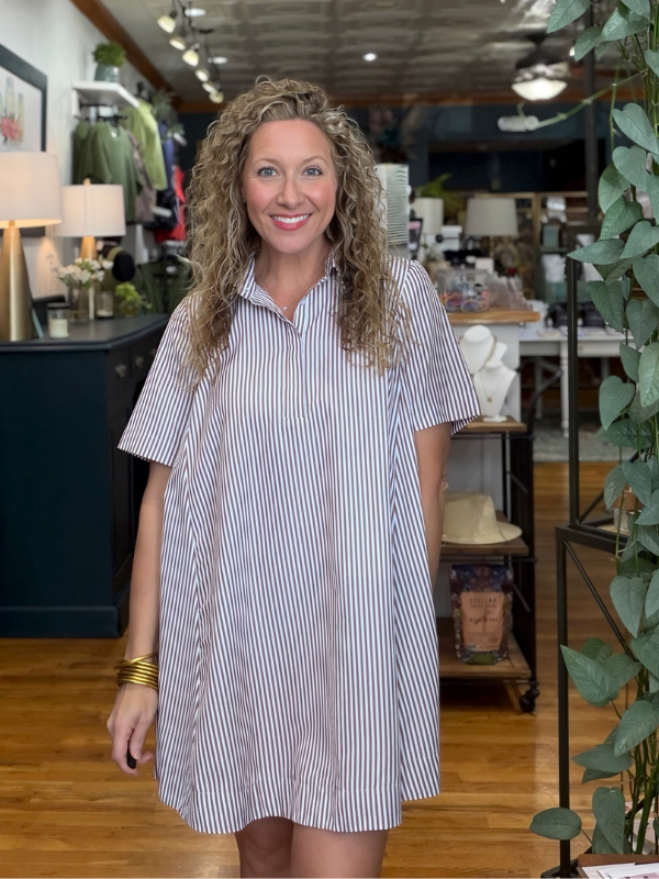 A smiling woman with curly blonde hair stands in a boutique wearing a short-sleeve, button-front shift dress with thin vertical blue and white stripes.