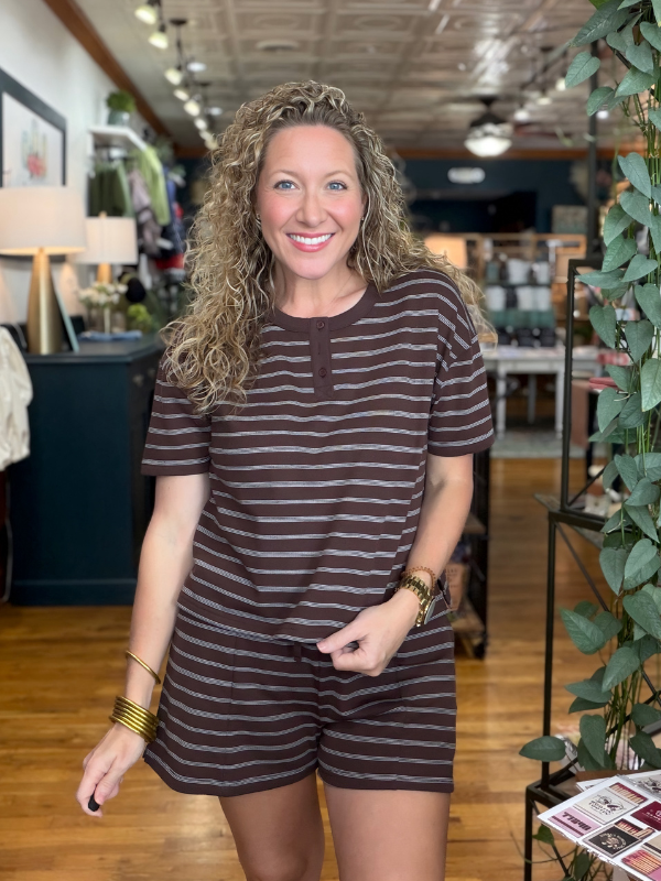 A smiling woman with curly blonde hair stands in a boutique wearing a short-sleeve brown and dark brown striped romper. The background shows a wooden floor, store fixtures, and a green plant.