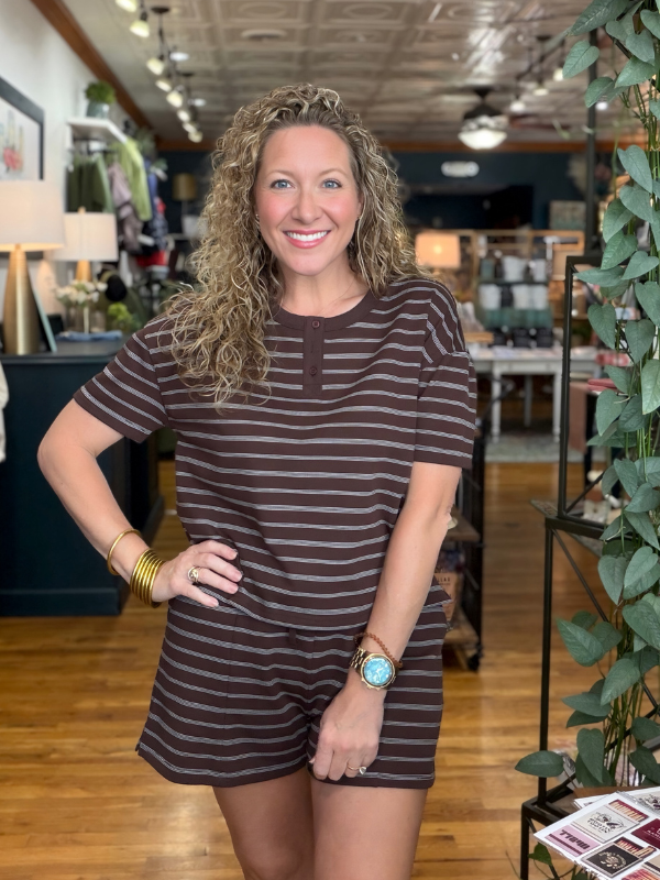 A smiling woman with curly blonde hair stands in a retail store wearing a brown and dark brown horizontally striped short-sleeve romper. She is wearing gold bracelets and a watch with a turquoise face.