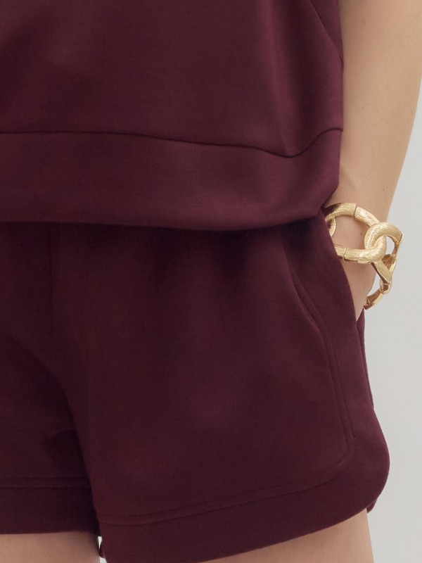 Burgundy shorts worn by a person with a gold bracelet on a light gray background