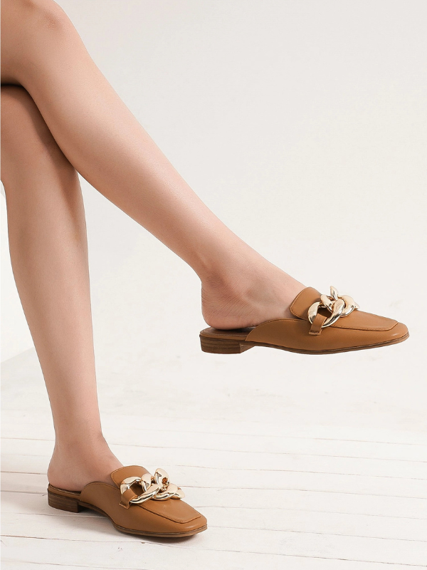 A pair of tan leather mule flats with chunky gold-tone chain embellishments on the strap, shown on a model's feet against a light off-white background.