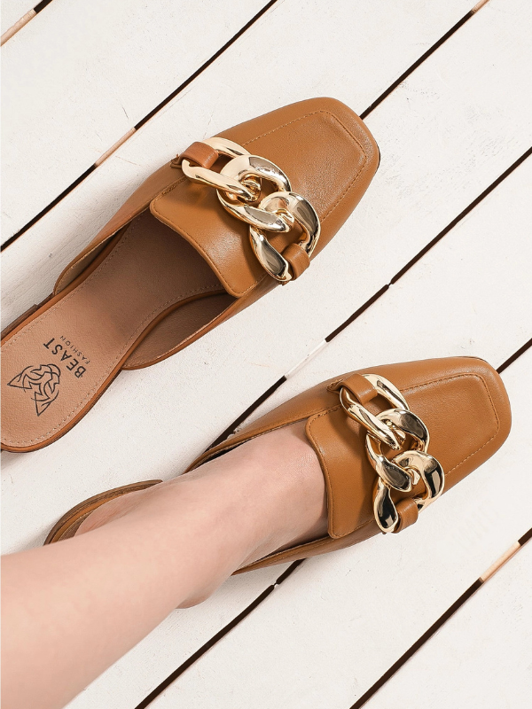 A pair of tan leather mule loafers with chunky gold chain embellishments, shown on a white wooden plank floor. One shoe is worn on a foot, and the brand name 'BEAST' is visible on the insole.