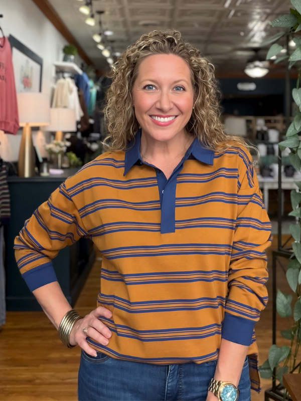 A smiling woman with curly blonde hair stands in a retail store wearing a mustard yellow and navy blue horizontally striped long-sleeve polo shirt with blue jeans.