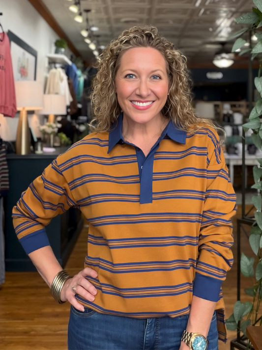 A smiling woman with curly blonde hair stands in a retail store wearing a mustard yellow and navy blue horizontally striped long-sleeve polo shirt with blue jeans.