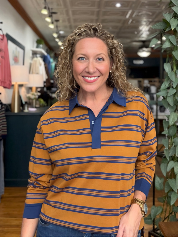 A smiling woman with curly blonde hair wearing a mustard yellow and navy blue striped long-sleeve polo shirt, standing inside a retail store.
