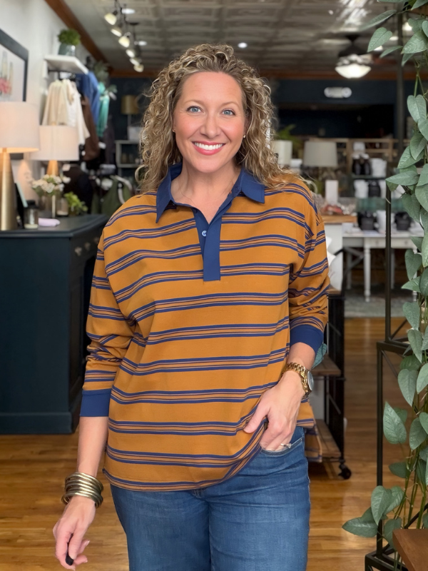 A smiling woman with curly blonde hair stands in a retail store wearing a long-sleeve polo shirt with horizontal mustard yellow and navy blue stripes, paired with blue jeans.