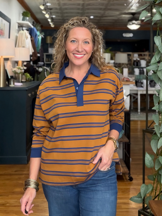 A smiling woman with curly blonde hair stands in a retail store wearing a long-sleeve polo shirt with horizontal mustard yellow and navy blue stripes, paired with blue jeans.