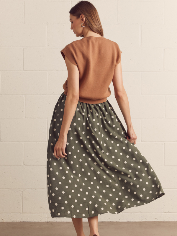 Woman wearing a brown top and green polka dot skirt against a beige tiled wall.