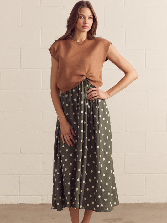 Woman wearing a brown top and polka dot skirt against a beige tiled wall.