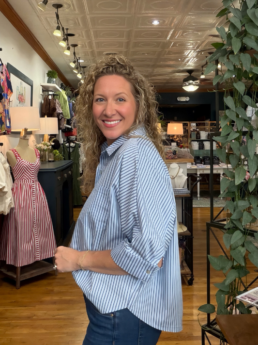 A smiling woman with curly blonde hair stands inside a boutique shop, wearing a light blue and white vertically striped button-down shirt with dark blue jeans.