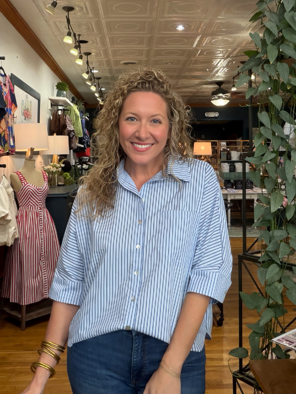 A smiling woman with curly blonde hair stands in a boutique wearing a light blue and white vertically striped button-down shirt with dark blue jeans.