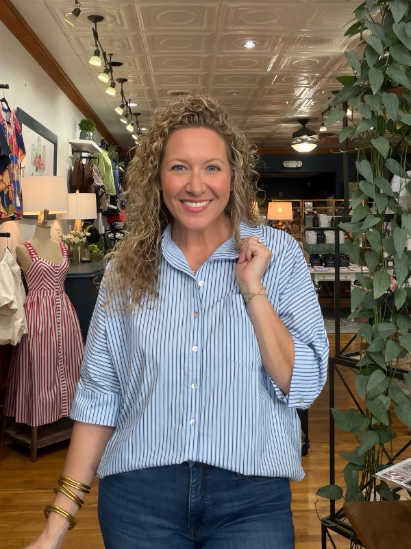 A smiling woman with curly blonde hair stands inside a clothing store wearing a light blue and white vertically striped button-down shirt with dark blue jeans.