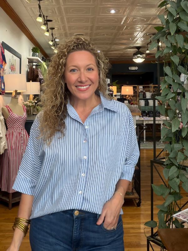 A smiling woman with curly blonde hair wearing a light blue and white vertically striped button-down shirt and dark blue jeans, standing inside a retail store.