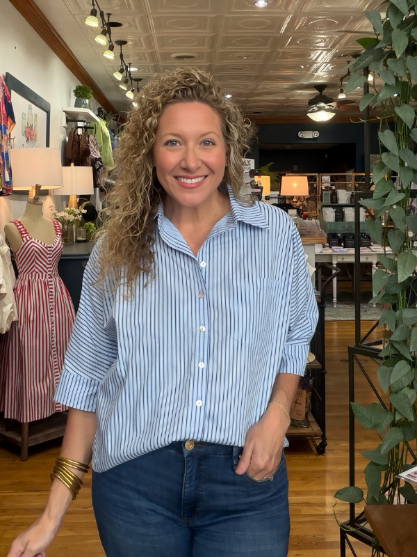 A smiling woman with curly hair wearing a light blue and white vertically striped button-down shirt and dark blue jeans, standing inside a retail store.