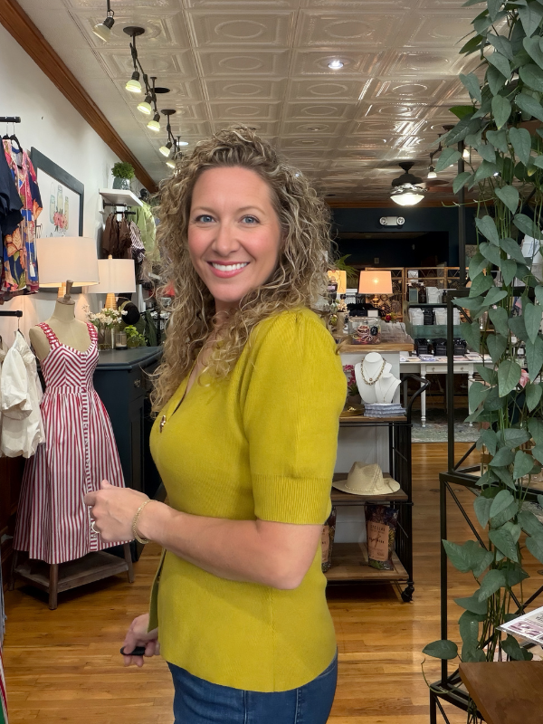 A smiling woman with curly blonde hair stands inside a boutique shop wearing a mustard yellow short-sleeve knit top and blue jeans. The shop has wood floors, a decorative ceiling, clothing racks, and a potted plant on the right.