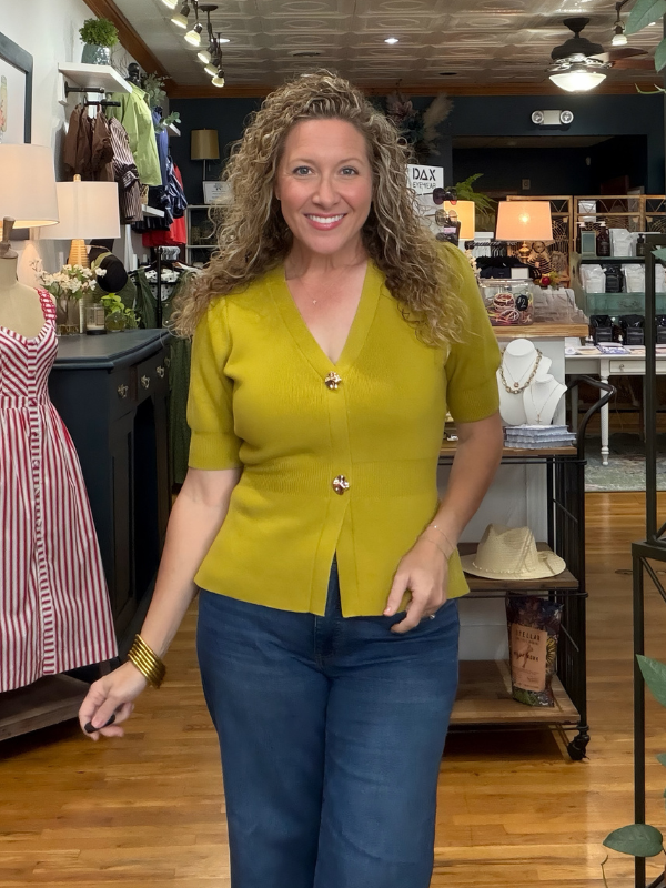 A smiling woman with curly hair stands in a boutique wearing a mustard yellow short-sleeve button-front top with blue jeans.