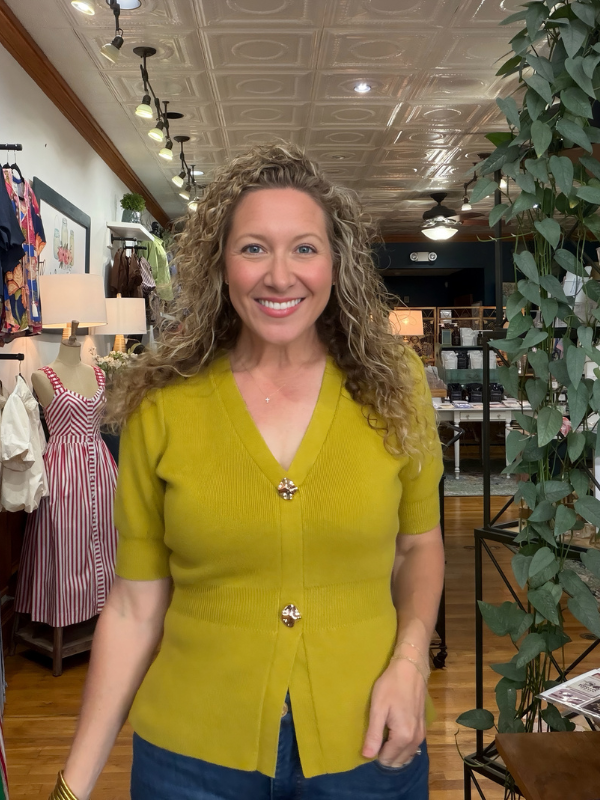 A smiling woman with curly blonde hair wearing a mustard yellow short-sleeve knit cardigan with decorative gold-tone buttons, standing inside a boutique store.