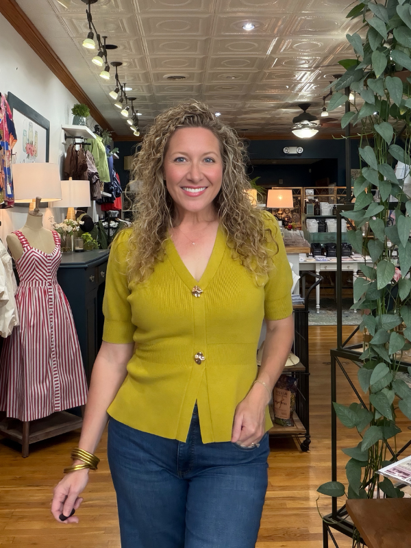 A smiling woman with curly blonde hair stands in a boutique shop, wearing a mustard yellow short-sleeve knit cardigan with gold buttons and blue jeans.