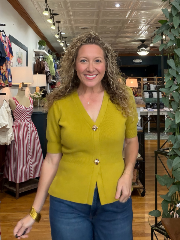 A smiling woman with curly hair wearing a mustard yellow short-sleeve button-front cardigan and blue jeans, standing inside a clothing boutique.