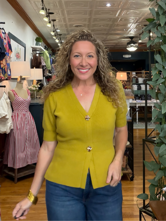 A smiling woman with curly hair wearing a mustard yellow short-sleeve button-front cardigan and blue jeans, standing inside a clothing boutique.