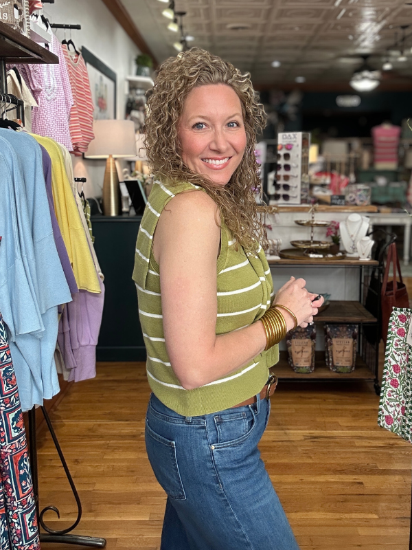 Woman in a green striped shirt and blue jeans standing in a store with clothing racks and products in the background.