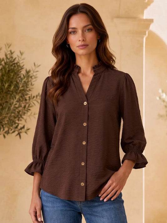 A woman wearing a dark brown crinkle-textured button-up blouse with ruffled collar and cuffs, paired with blue jeans, standing against a warm beige architectural background.