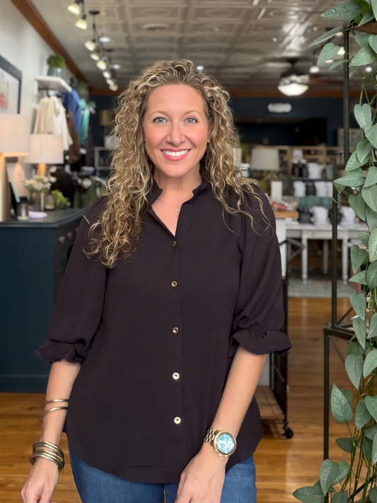 A smiling woman with curly blonde hair wearing a dark brown button-down shirt and blue jeans, standing in a retail store interior.