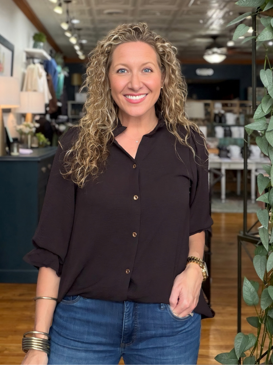 A smiling woman with curly blonde hair stands in a boutique wearing a dark brown button-down blouse and blue jeans.
