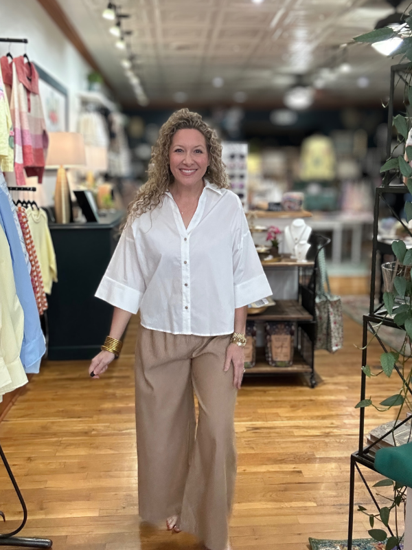 Woman in a white blouse and beige pants standing in a store with clothing racks and displays.