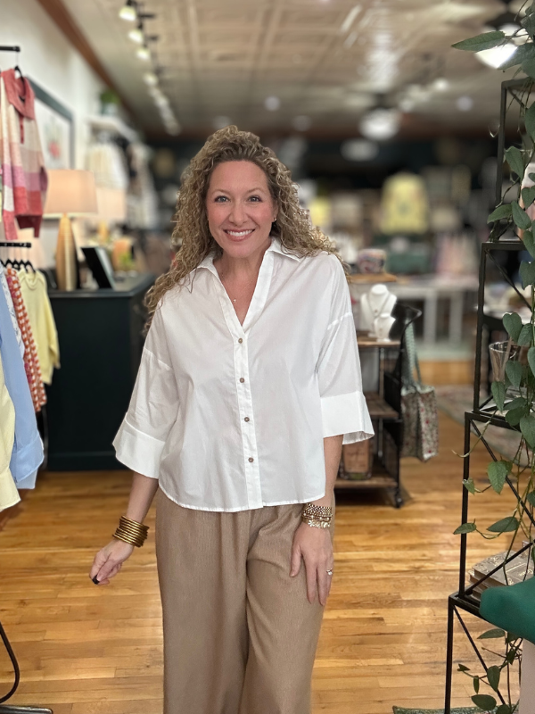Woman in a white blouse and beige pants standing in a store with wooden floors and clothing racks.