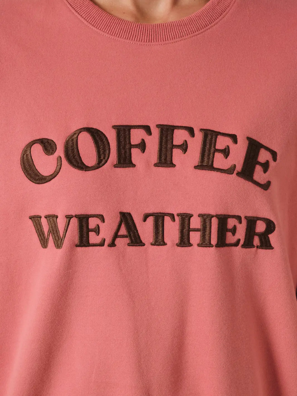 A close-up of a dusty pink crewneck sweatshirt featuring the words 'COFFEE WEATHER' embroidered in dark brown across the chest.