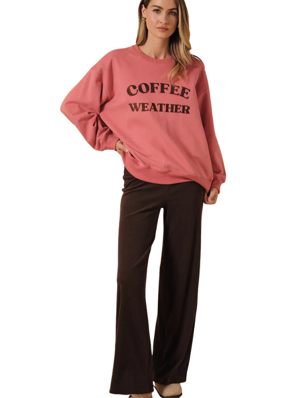 A woman modeling a dusty pink crewneck sweatshirt with 'COFFEE WEATHER' printed in black across the chest, paired with dark brown wide-leg pants, against a plain white background.