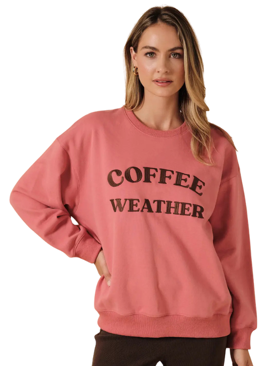 A woman modeling a dusty rose crewneck sweatshirt with 'COFFEE WEATHER' printed in black arched text across the chest, set against a plain white background.