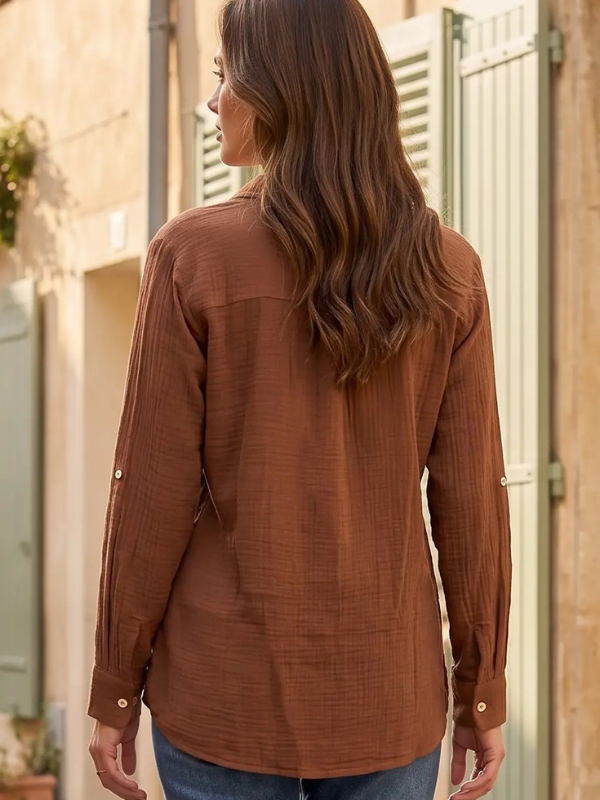 A woman viewed from behind wearing a brown textured long-sleeve shirt and blue jeans, standing outdoors in front of a beige building with white shutters.