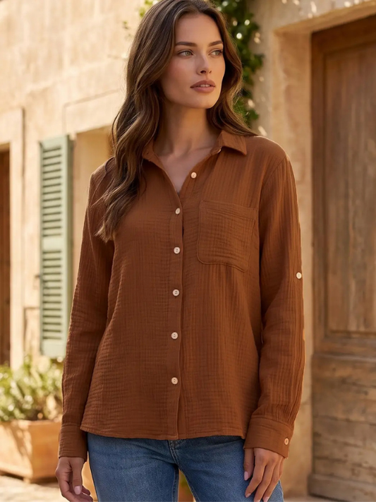 A woman wearing a textured brown long-sleeve button-up shirt with a chest pocket, paired with blue jeans, standing outdoors in front of a rustic stone building.