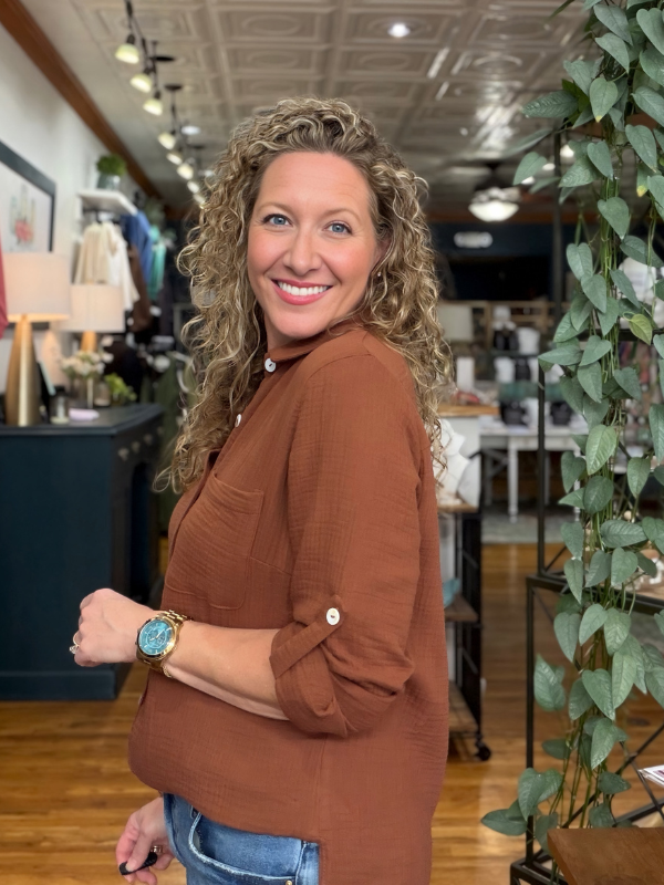 A smiling woman with curly blonde hair stands in a boutique wearing a rust-brown long-sleeve button-up shirt and light-wash denim shorts.