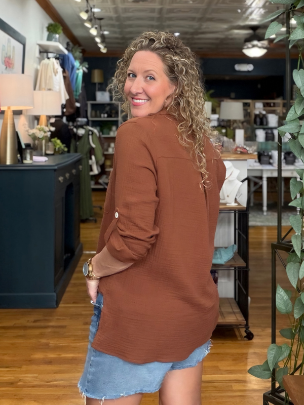 A woman with curly blonde hair wearing a rust-brown long-sleeve button-up shirt and denim shorts, standing in a retail store and looking back over her shoulder.
