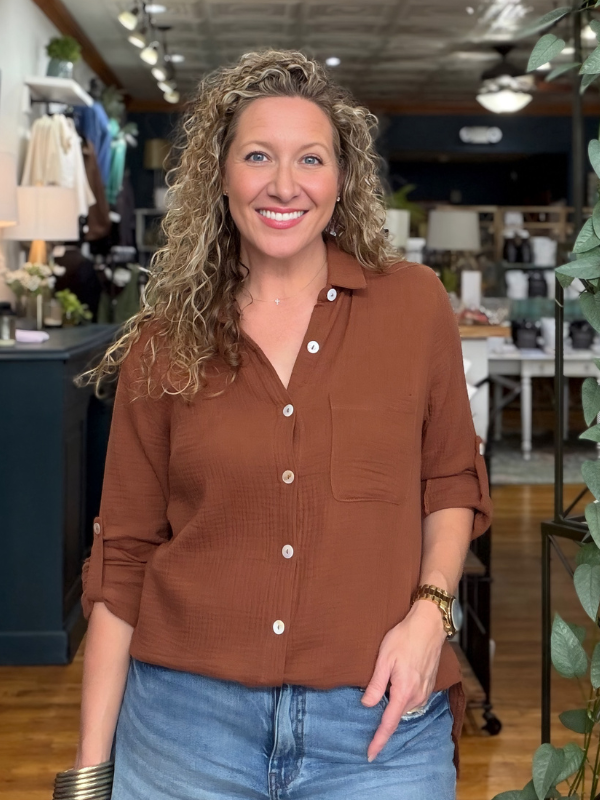 A smiling woman with curly blonde hair stands in a boutique wearing a rust-brown long-sleeve button-up shirt and light-wash denim shorts.