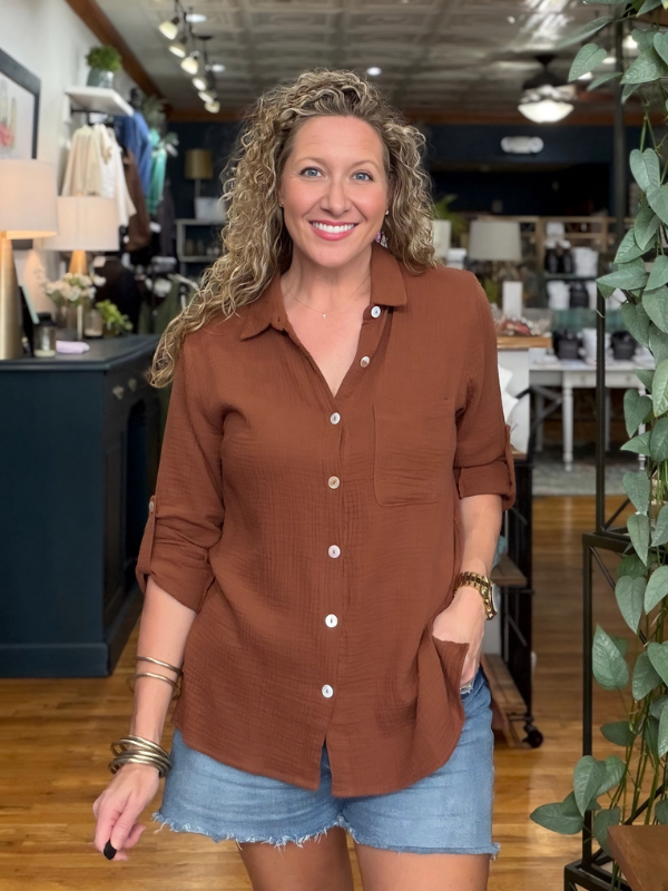 A smiling woman with curly blonde hair stands in a boutique wearing a rust-brown long-sleeve button-up shirt and light-wash denim shorts.