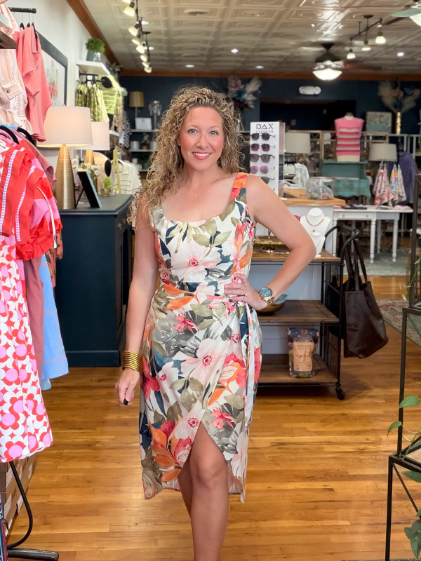 Woman in a floral dress standing in a store with clothing racks and products in the background.