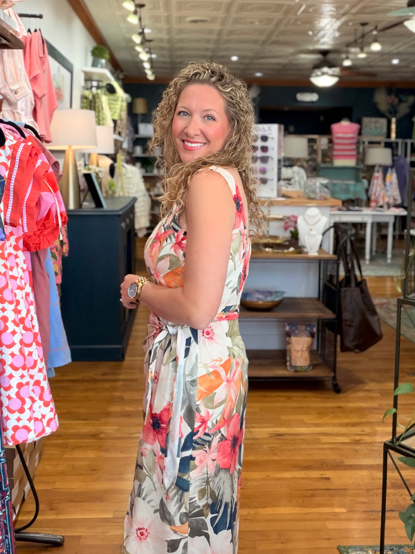 Woman in a floral dress standing in a store with clothing racks and products in the background.