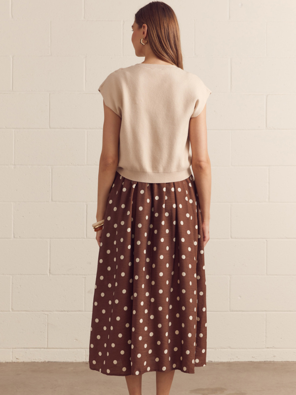 Woman wearing a beige top and brown polka dot skirt against a white brick wall.