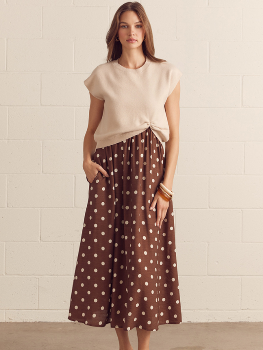 Woman wearing a beige top and brown polka dot skirt against a light brick wall.